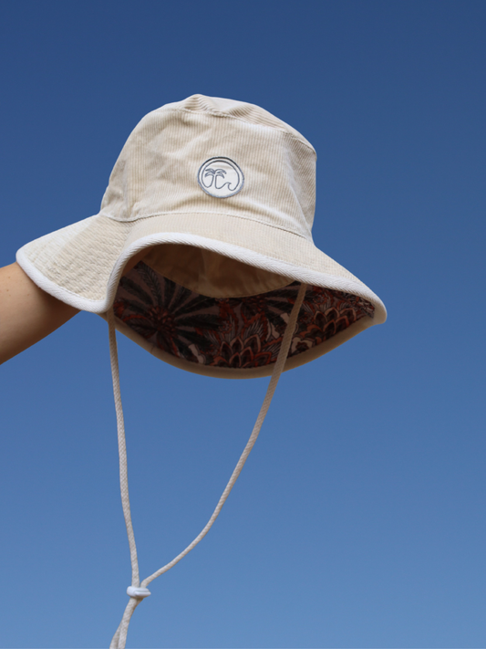 Beige corduroy bucket hat with a morals the brand logo against a clear blue sky